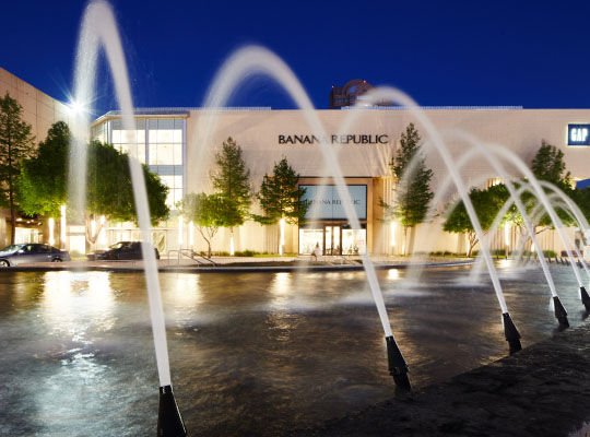 Night view of a shopping mall entrance with water fountains arcing in front of the Banana Republic store, illuminated by warm lights.