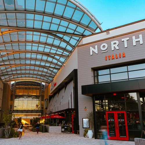 Exterior view of North Italia restaurant with a modern glass-covered walkway and people walking by, taken at twilight.