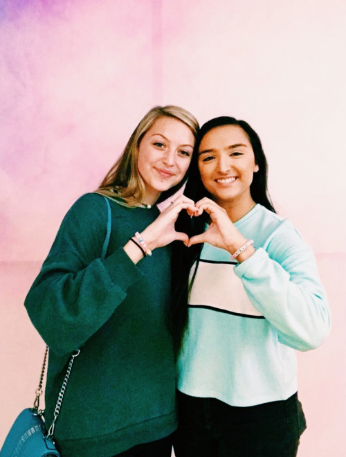 Two young women smiling and making a heart shape with their hands, standing in front of a pink and purple gradient background.