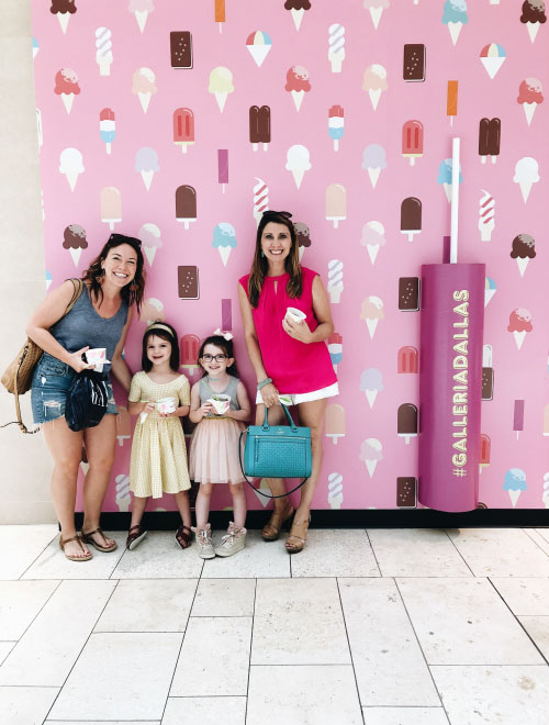 Two women and two young girls posing in front of a pink wall decorated with ice cream illustrations at Galleria Dallas, holding cups of ice cream and smiling.