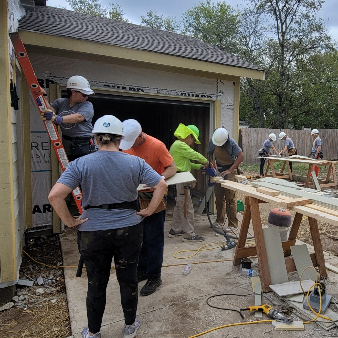 Group of volunteers wearing hard hats working on a house construction project. Some are using tools, while others are holding materials or working on a ladder.