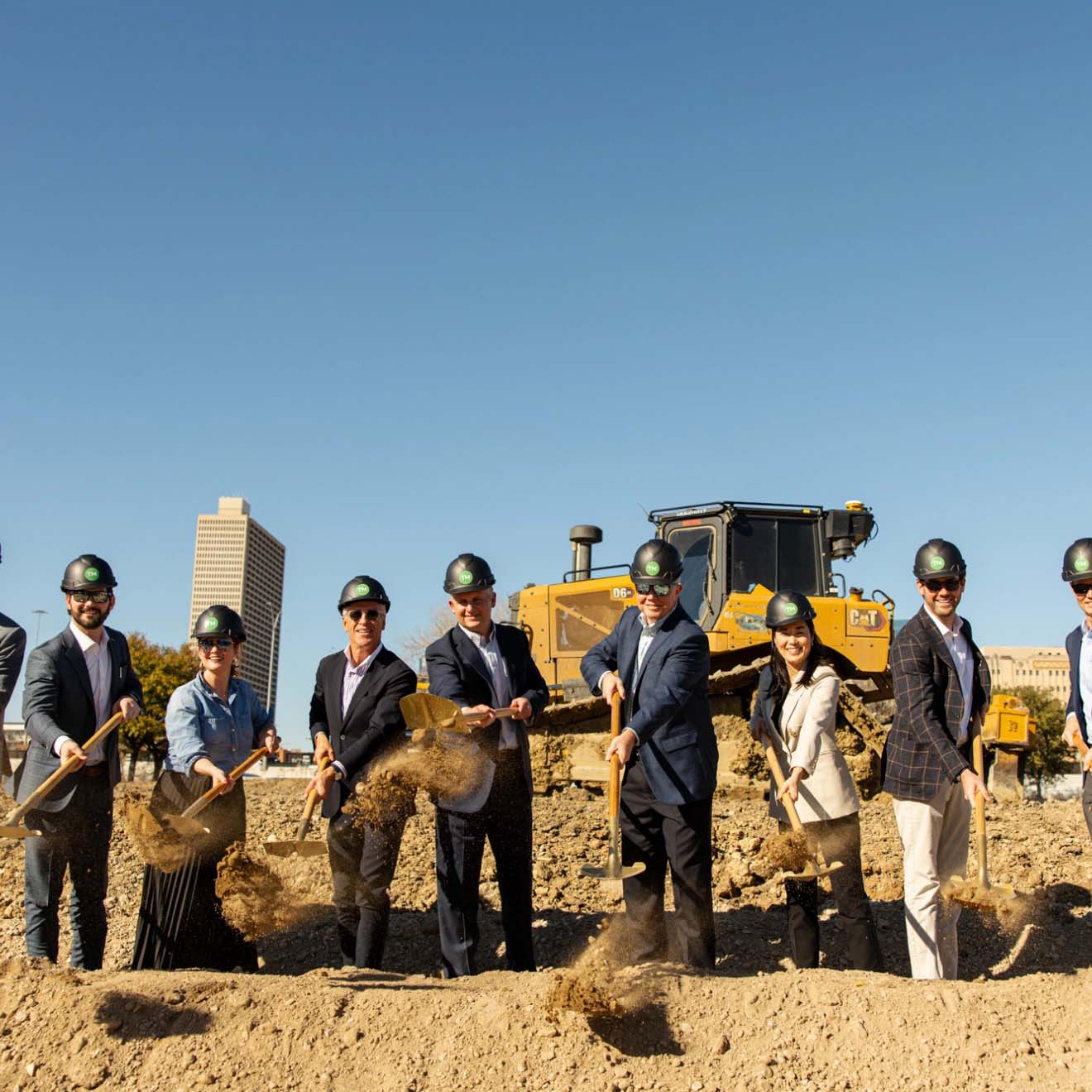 Group of people wearing hard hats and holding shovels at a groundbreaking ceremony, with a bulldozer and city buildings in the background.