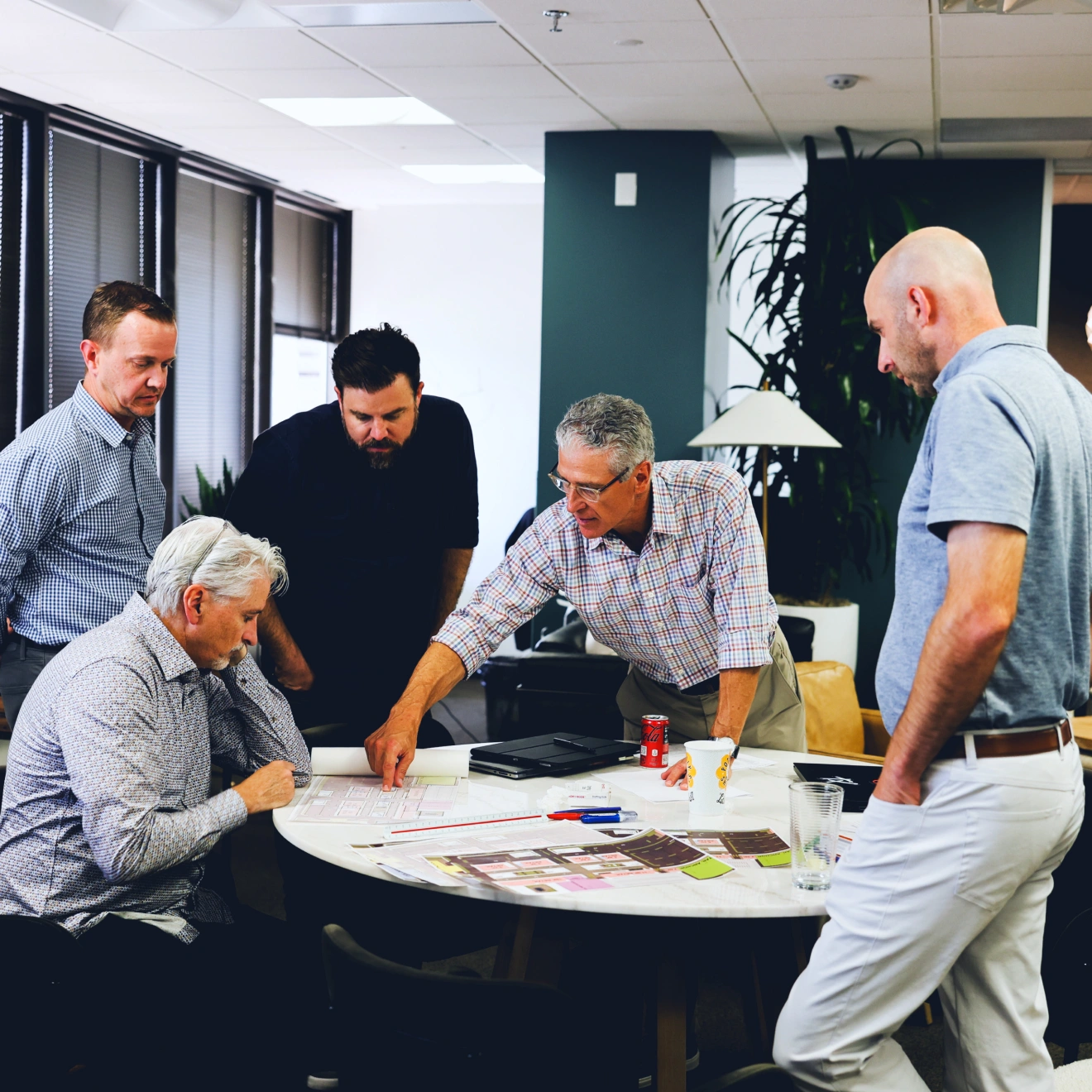 Group of five men gathered around a table in an office setting, discussing documents and plans. One man is pointing at a paper on the table, while the others look on attentively.