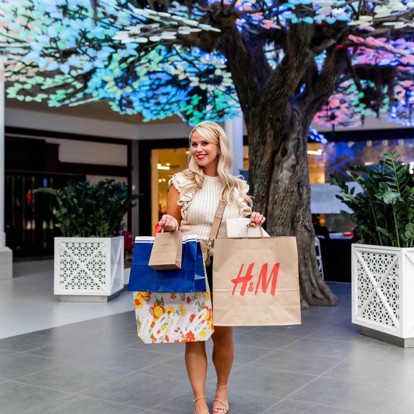 Woman smiling while holding several shopping bags from different stores, standing inside a mall with a colorful, illuminated tree sculpture in the background.