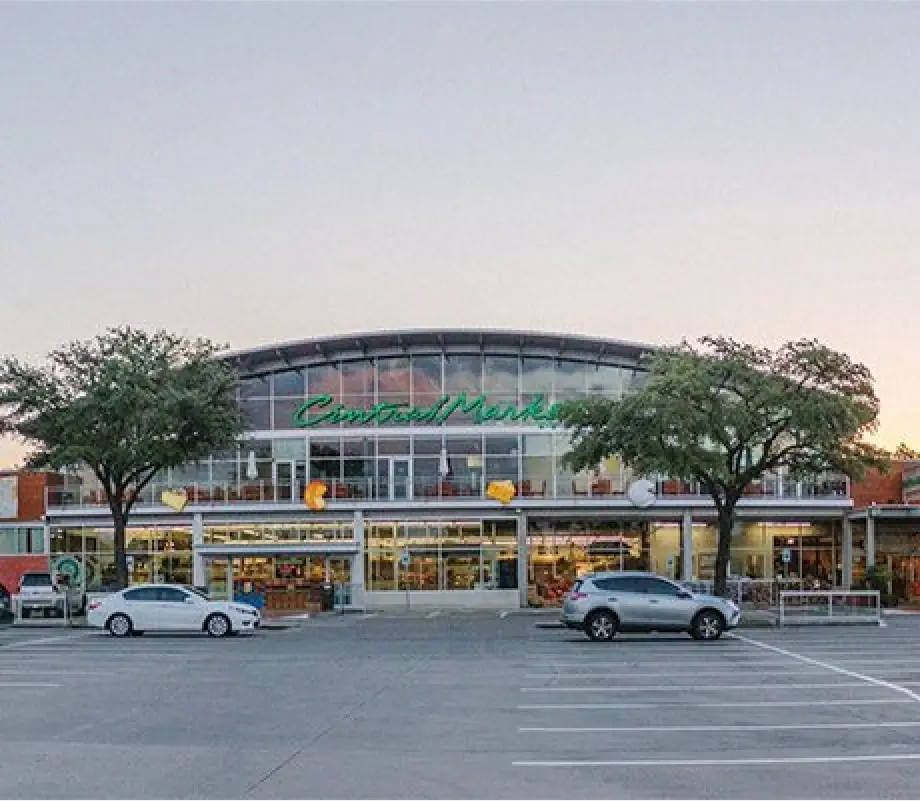 Front view of a Central Market store with a large glass facade, trees, and a parking lot in the foreground during early evening.