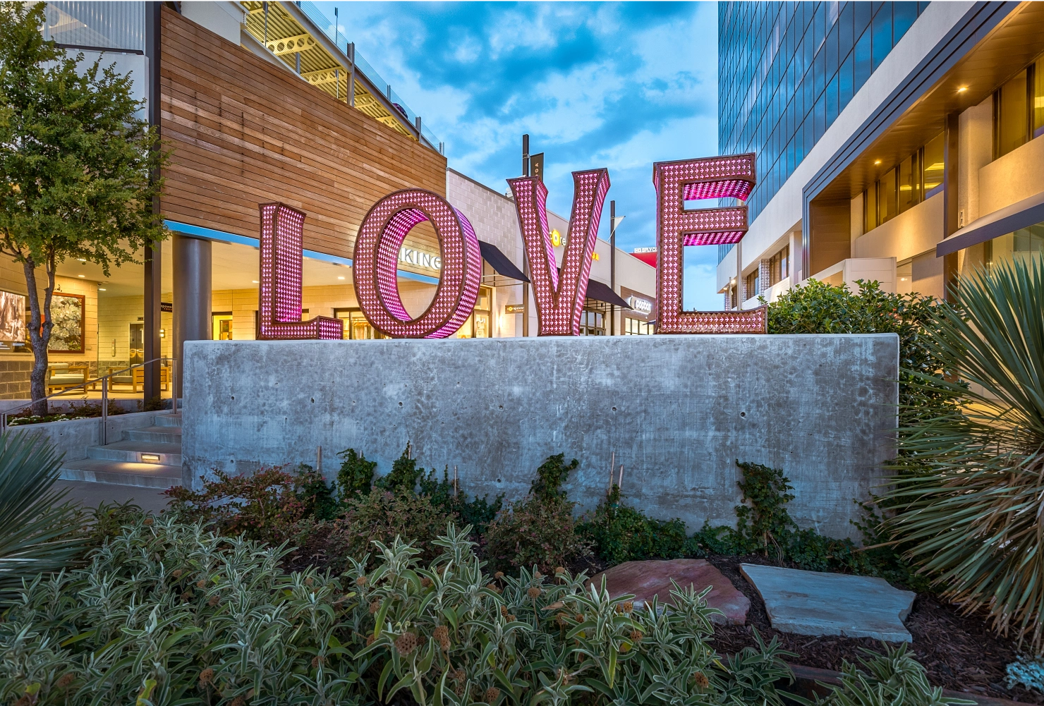 Large illuminated 'LOVE' sign in front of a modern shopping center with landscaping, taken during early evening.