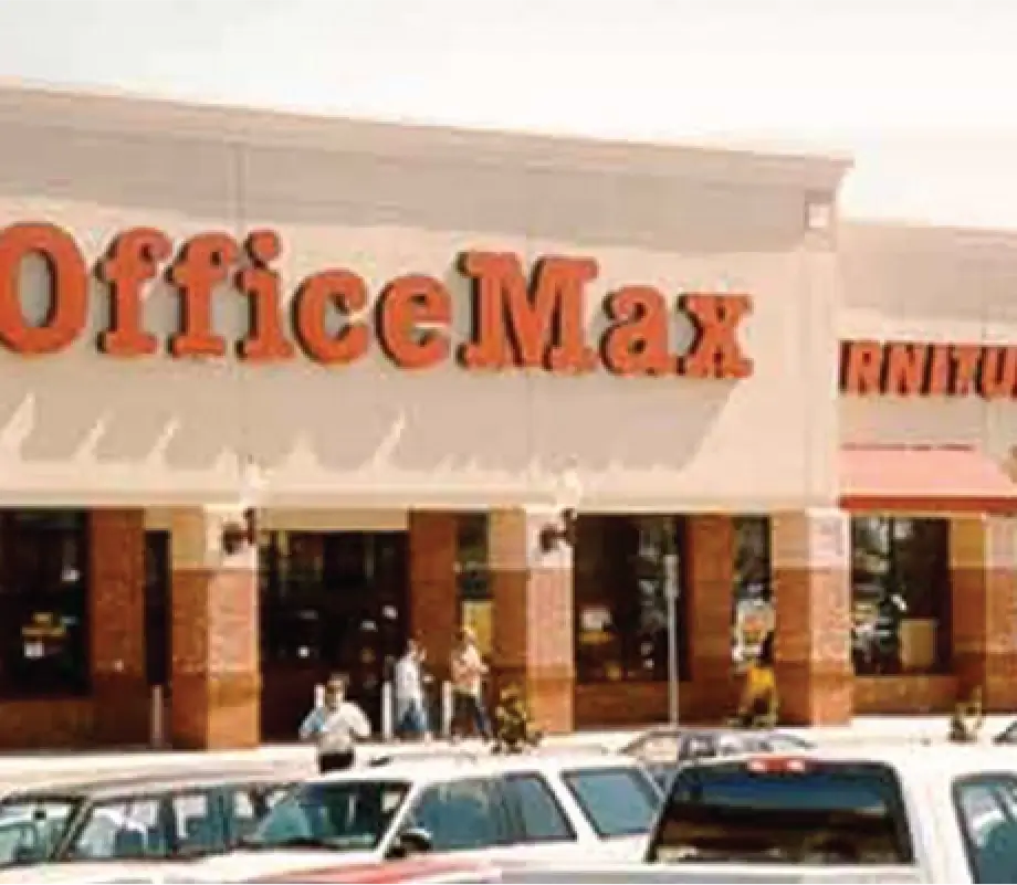 Exterior view of an OfficeMax store with large red signage, with people walking near the entrance and cars parked in front.