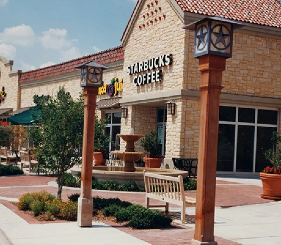 Exterior view of a Starbucks Coffee shop in a shopping center with stone architecture, surrounded by landscaping, benches, and wooden posts with lanterns.