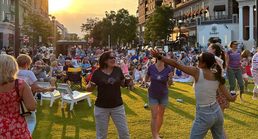 Outdoor community event at sunset with a large group of people gathered on a lawn, some dancing and others sitting in chairs, enjoying the lively atmosphere.